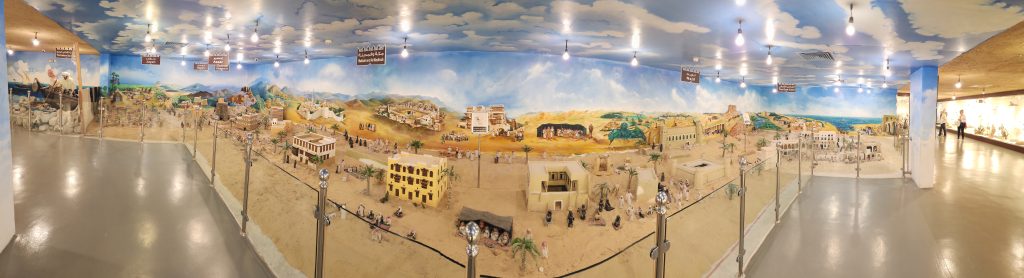 Panoramic view of a museum exhibit showcasing Arabian history, featuring miniature models of traditional buildings and desert landscapes, with vibrant murals depicting the region's heritage.