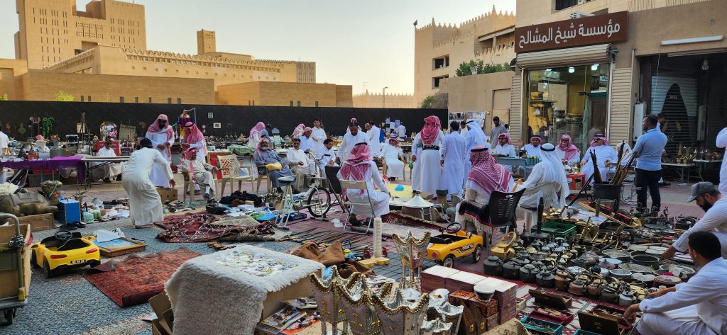 Bustling market scene in Riyadh with traders in traditional attire, showcasing antiques and vintage items amidst vibrant displays of goods.