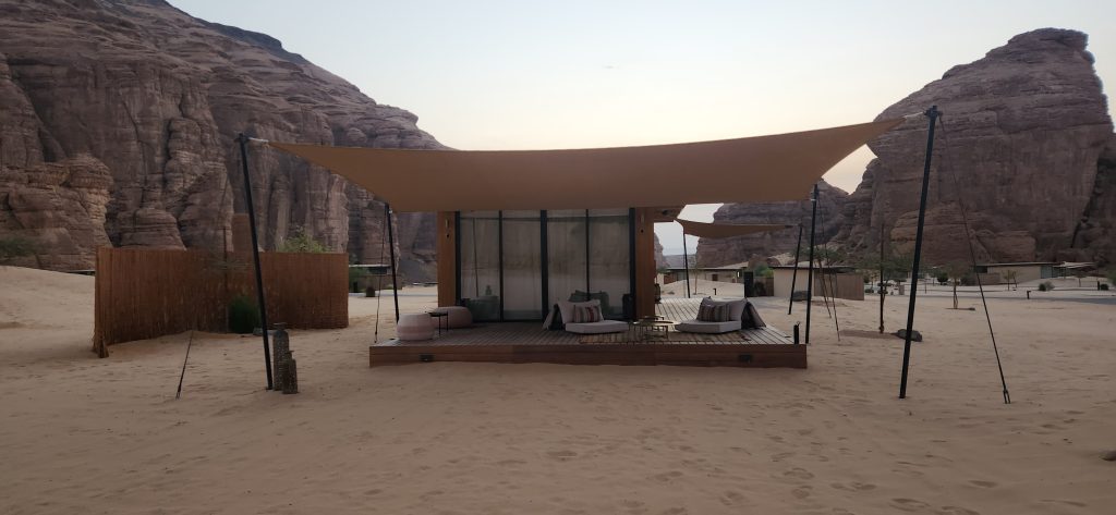 Desert lodge with modern architecture and shaded seating area, framed by sandstone cliffs in AlUla, Saudi Arabia.