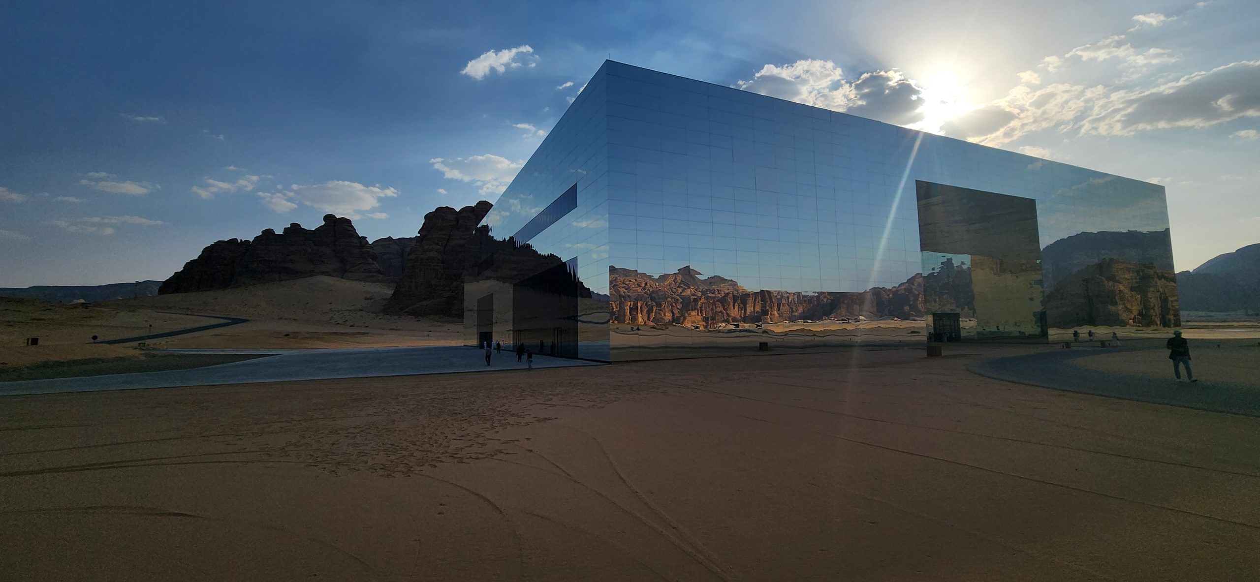Modern reflective building in desert landscape, showcasing Nabataean tombs and rock formations, under a blue sky with clouds, highlighting Saudi Arabia's unique architecture and natural beauty.