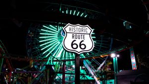 Historic Route 66 sign illuminated at night with a colorful ferris wheel in the background, symbolizing adventure and unique travel experiences.
